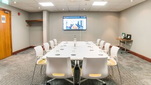 Une salle de conférence avec une longue table, des chaises blanches, un écran de projection et une petite table sur laquelle se trouvent un téléphone et une boîte.