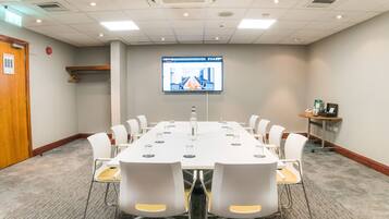 A conference room with a long table, white chairs, a projector screen, and a small table with a phone and a box.