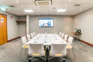 Une salle de conférence avec une longue table, des chaises blanches, un écran de projection et une petite table sur laquelle se trouvent un téléphone et une boîte.