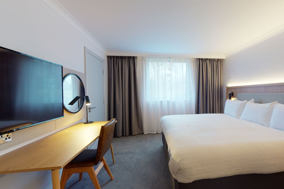 Une chambre d’hôtel comprenant un lit, un bureau avec une chaise, un téléviseur à écran plat, une grande fenêtre avec des rideaux et une tête de lit dotée d’un luminaire.