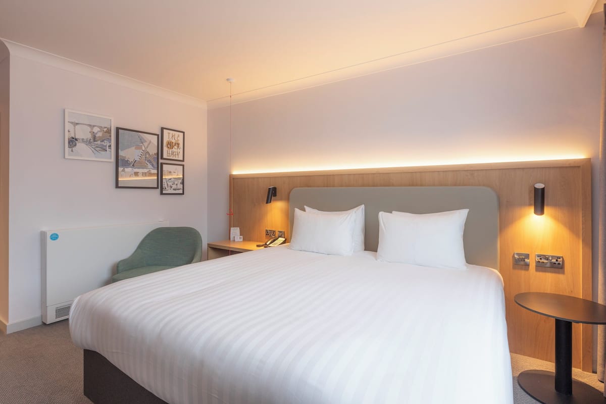 Une chambre d’hôtel avec un grand lit, une table de chevet avec une lampe, une chaise et des tableaux encadrés au mur.