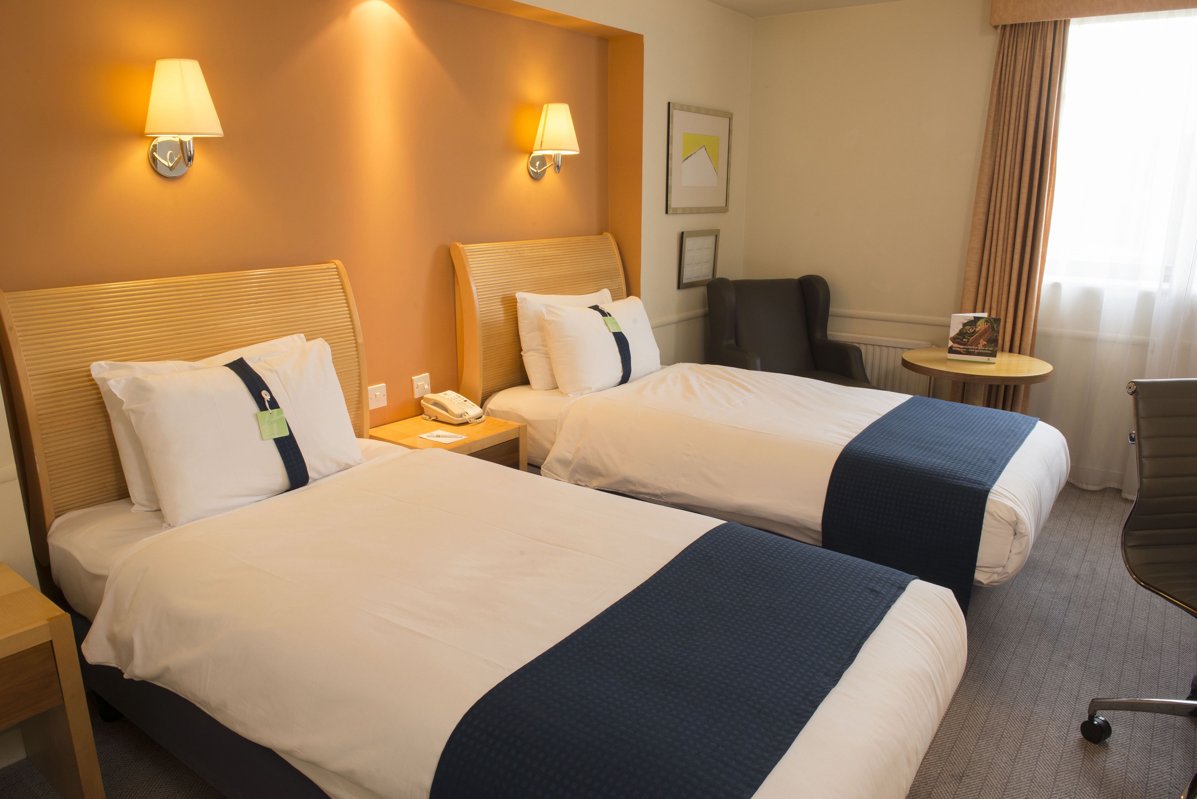 Habitació d'hotel amb dos llits, cadascun amb capçalera, taules de nit i làmpades. Hi ha una taula rodona petita i una cadira a l'habitació.
