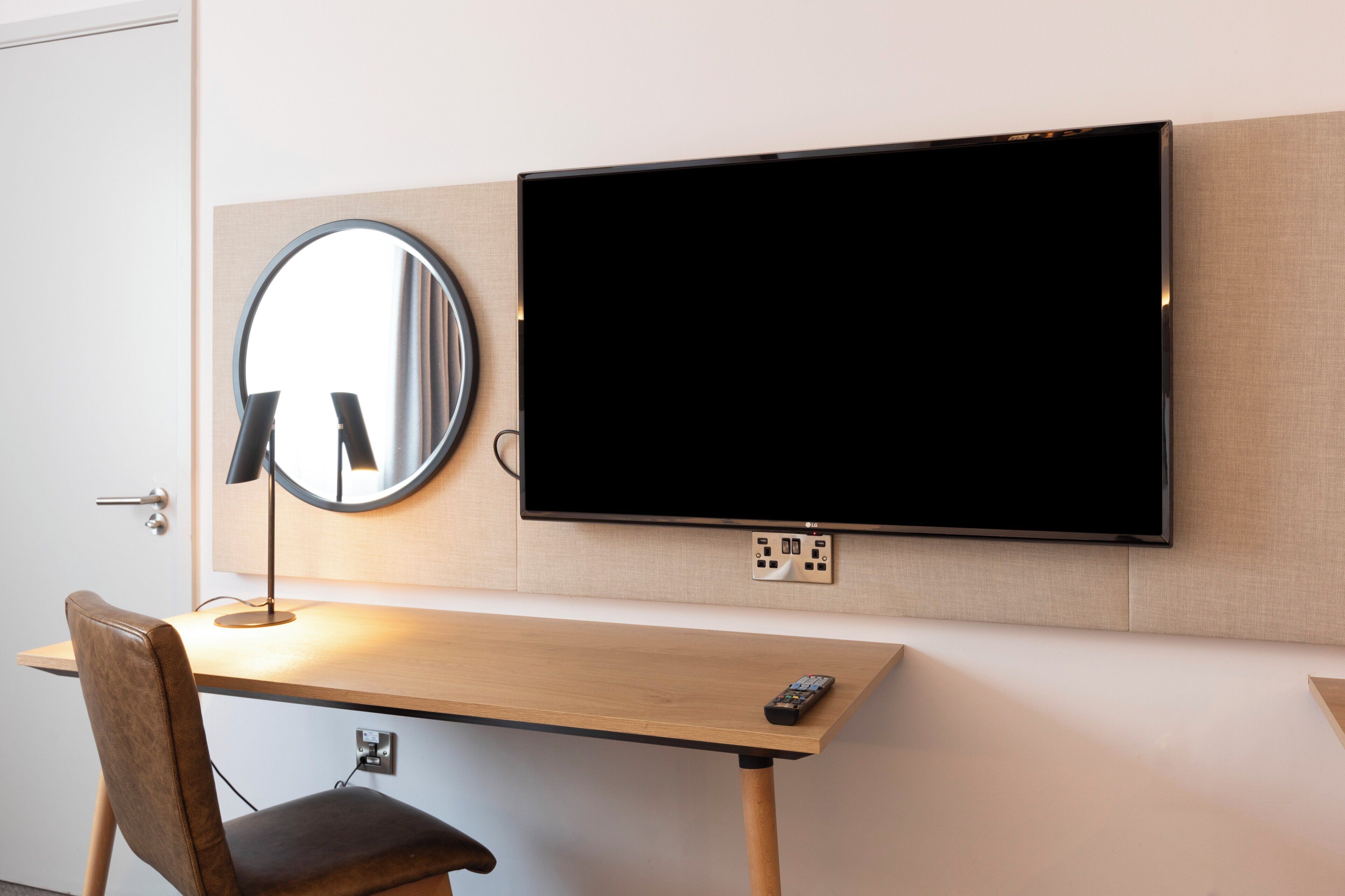 Habitación de hotel con televisor de pantalla plana montado en la pared, un espejo redondo, un escritorio de madera con una silla y un control remoto sobre el escritorio.