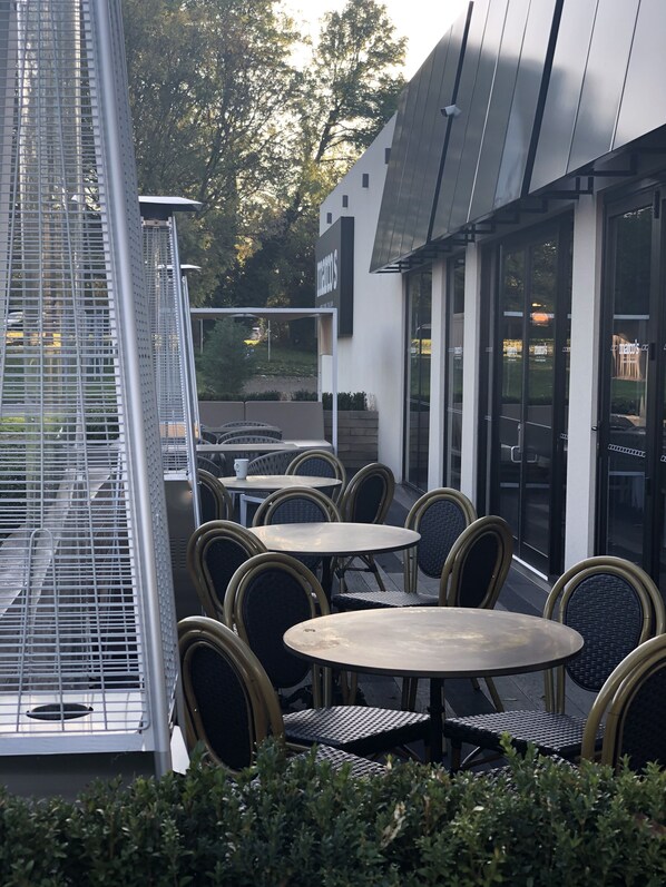 Un espace extérieur aménagé avec des tables et des chaises rondes, une structure de barbecue en métal et un bâtiment sur lequel on peut lire l’inscription « MEO’S ».