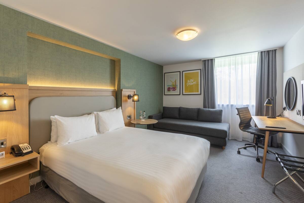 Une chambre d’hôtel avec un grand lit, un bureau avec une chaise, un canapé, une petite table avec une lampe et un téléphone sur une table de chevet.
