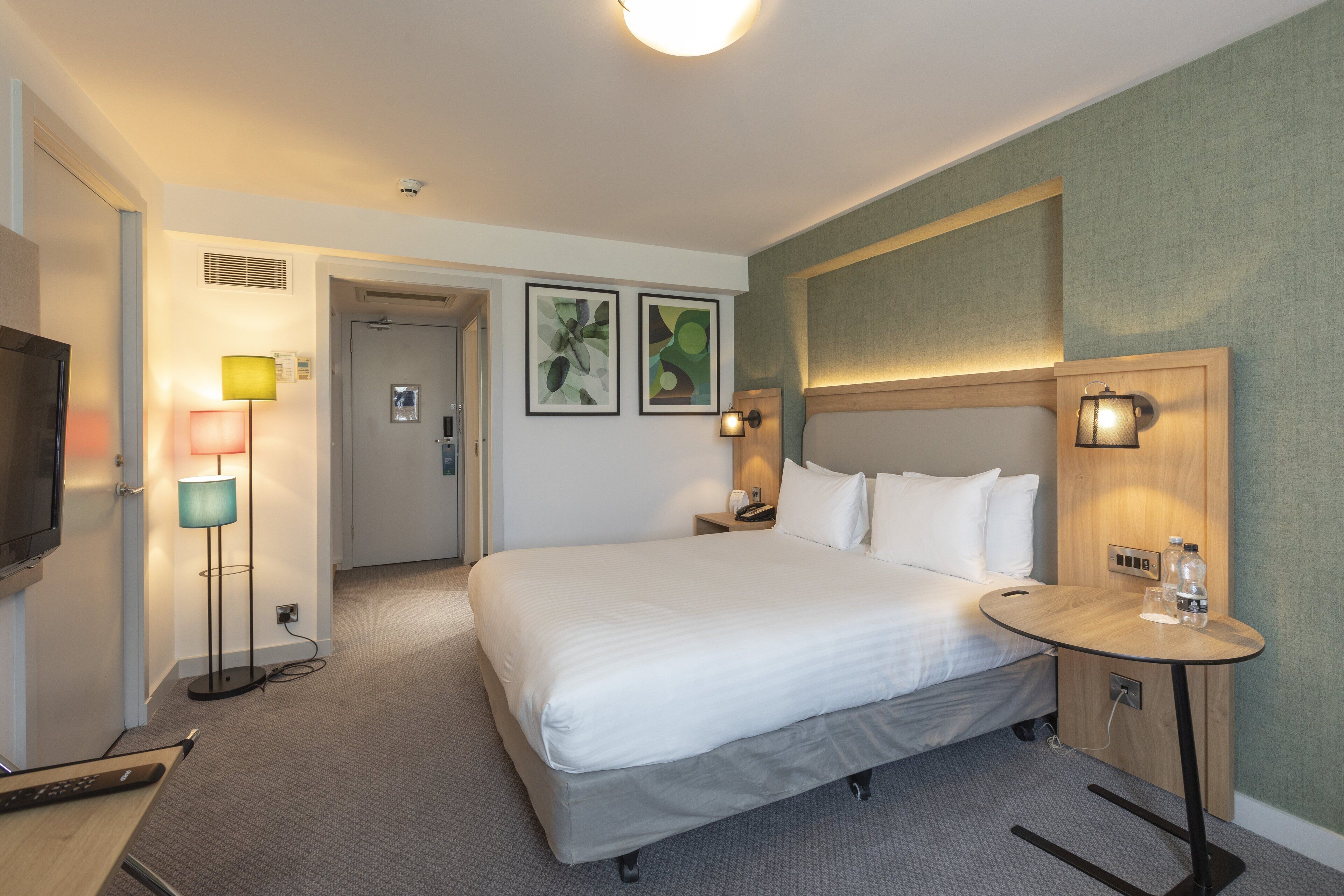Een hotelkamer met een bed, nachtkastje, lamp en kunstwerk aan de wand.