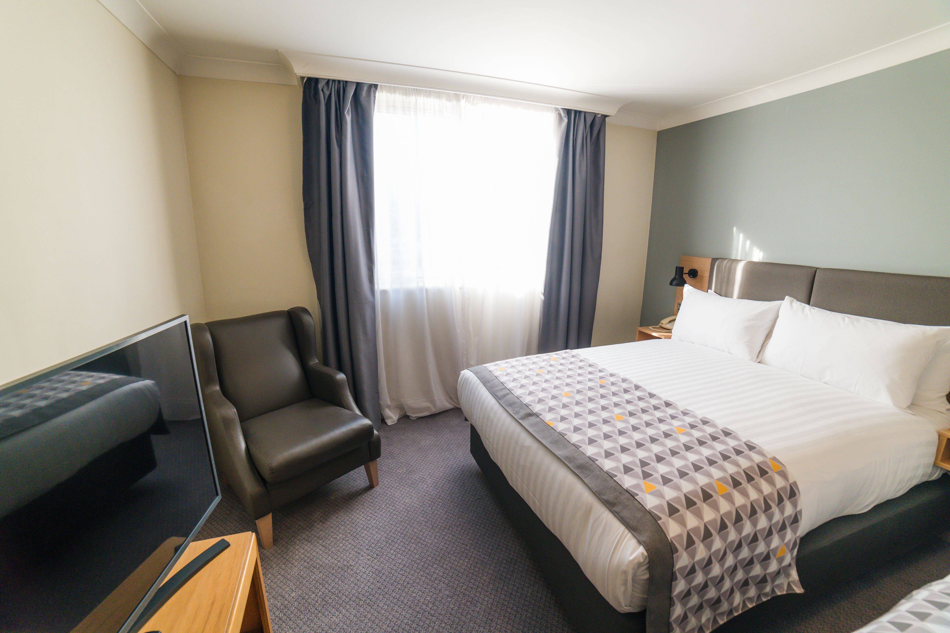 A hotel room with a bed, a chair, a desk, a TV, and a window with curtains.