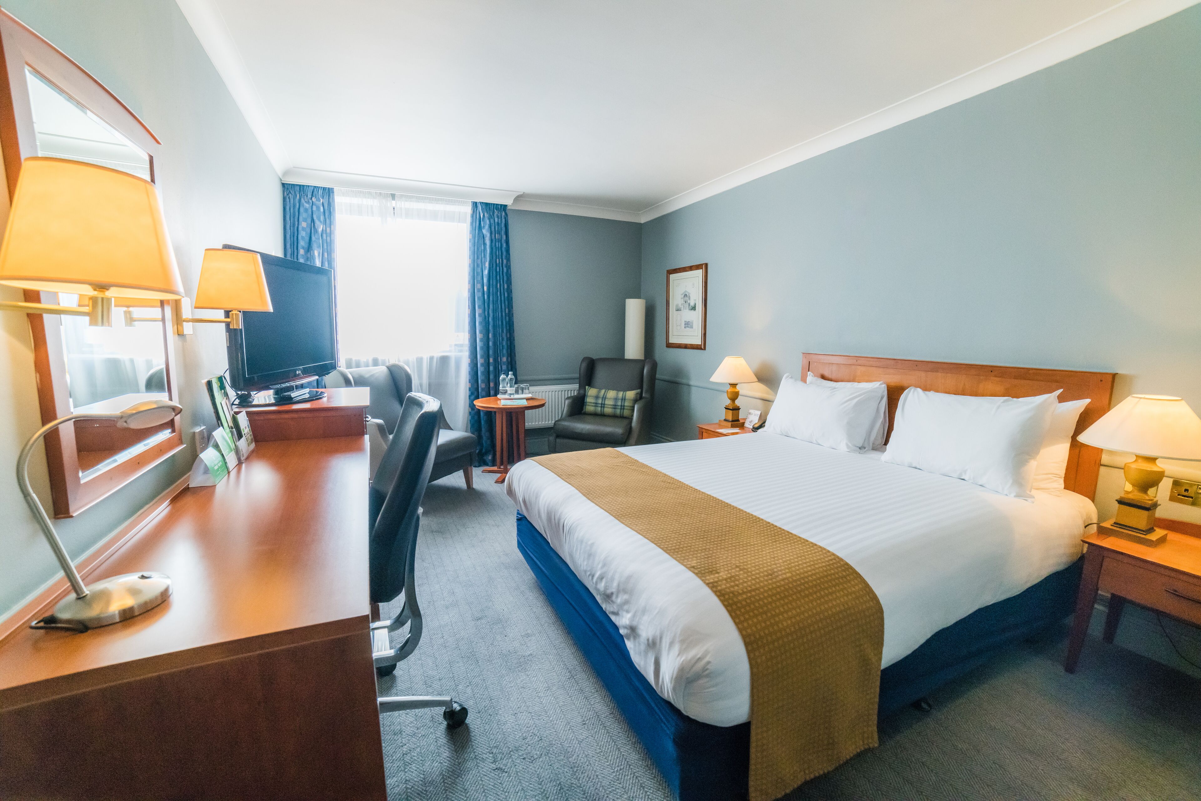 A hotel room with a large bed, a desk with a computer, a chair, a television, and a window with curtains.
