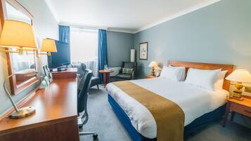 A hotel room with a large bed, a desk with a computer, a chair, a television, and a window with curtains.