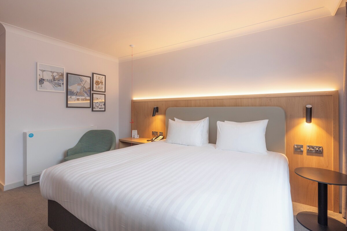 Une chambre d’hôtel avec un grand lit, une table de chevet avec une lampe, une chaise et des tableaux encadrés au mur.