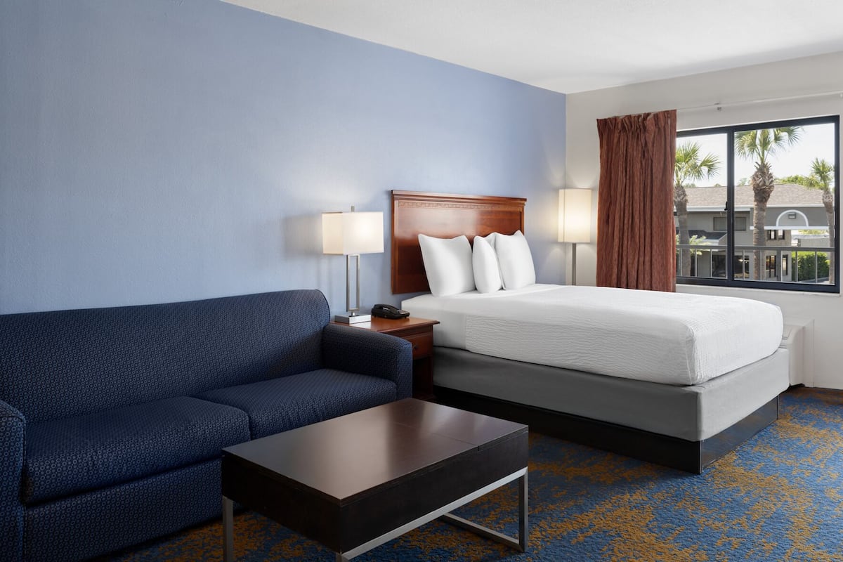 Une chambre d’hôtel avec un lit, un canapé, une table de chevet, une lampe et une fenêtre donnant sur des palmiers.