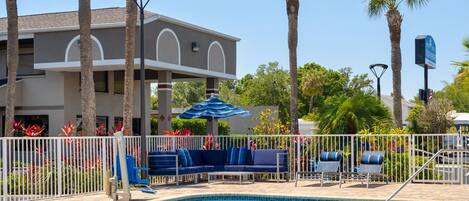 A pool area with lounge chairs and umbrellas, surrounded by palm trees and a building with a grey facade.