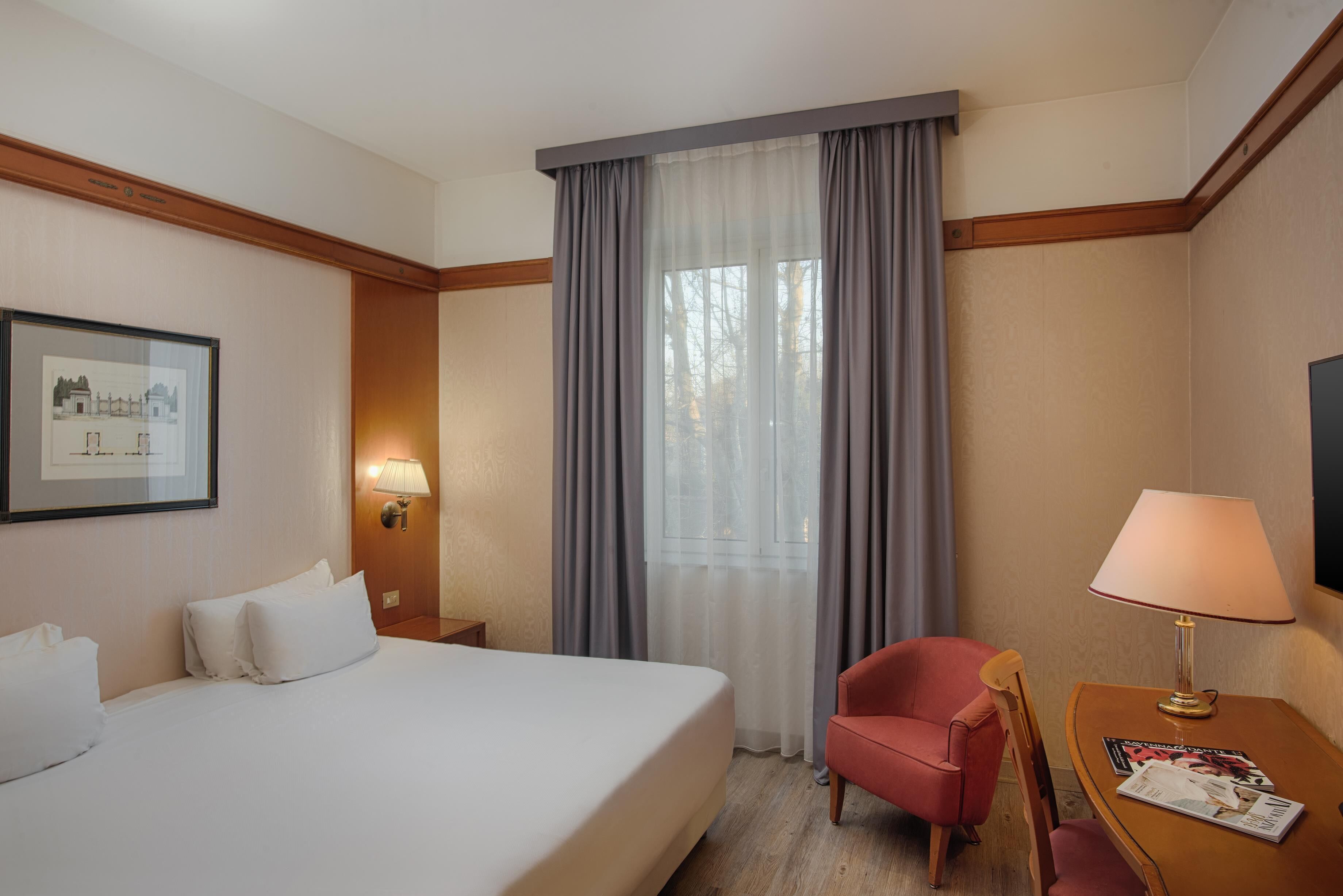 A hotel room with a large bed, a red chair, a wooden nightstand with a lamp, and a framed picture on the wall.