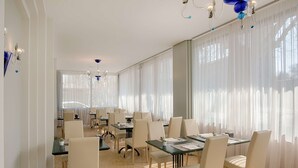 A conference room with multiple tables and chairs, large windows with sheer curtains, and a chandelier.
