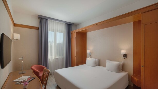 A hotel room with a bed, a desk with a chair, a TV, and a window with curtains.