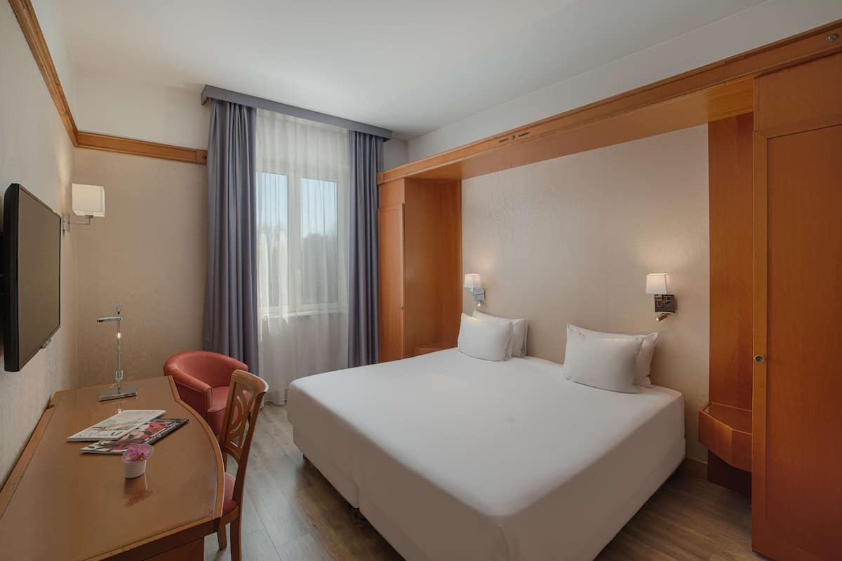 A hotel room with a bed, a desk with a chair, a TV, and a window with curtains.
