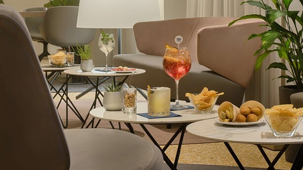 Un área de descanso moderna con asientos, una mesa con aperitivos y bebidas, y una planta en maceta.