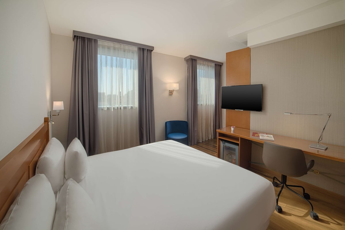 A hotel room with a large bed, a desk with a chair, a television, and a window with curtains.