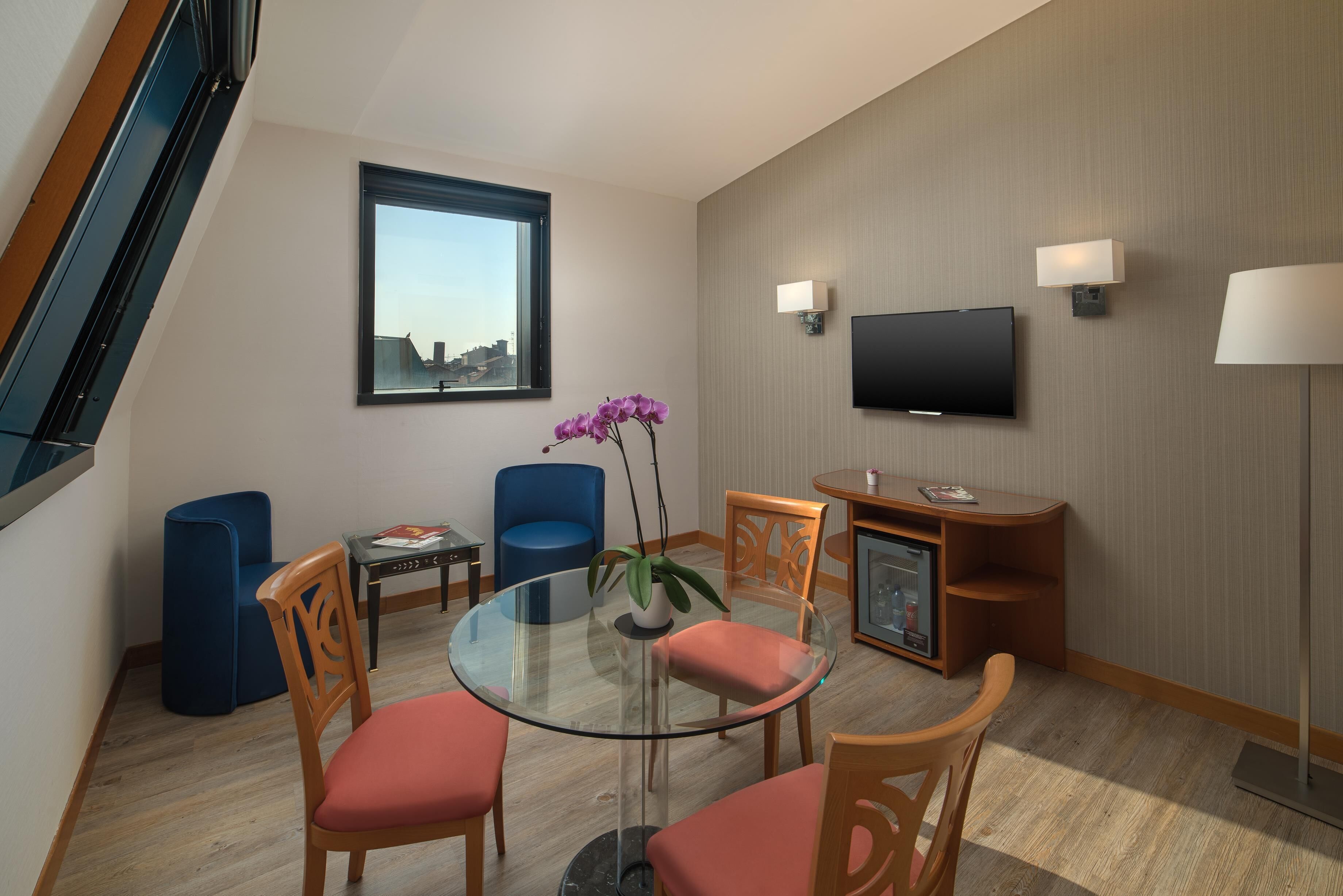 Ein modernes Hotelzimmer mit einem Glastisch, Holzstühlen, einem blauen Sofa, einem kleinen Beistelltisch, einem an der Wand montierten Fernseher und einem Fenster mit Blick auf Gebäude.