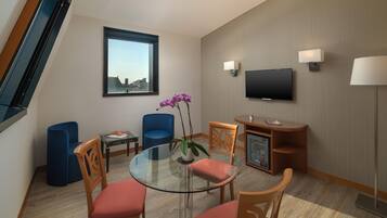 Ein modernes Hotelzimmer mit einem Glastisch, Holzstühlen, einem blauen Sofa, einem kleinen Beistelltisch, einem an der Wand montierten Fernseher und einem Fenster mit Blick auf Gebäude.