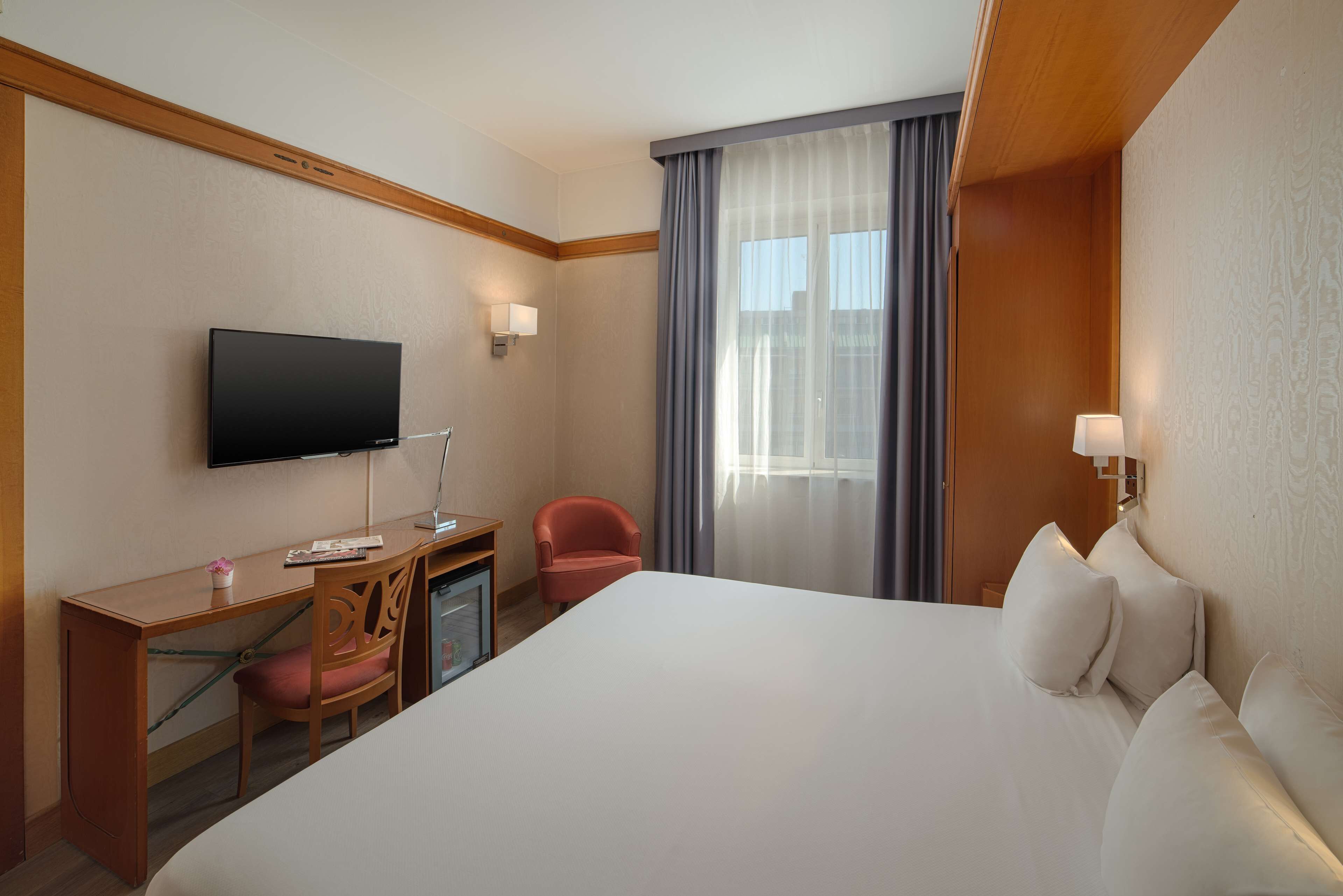 Ein Hotelzimmer mit einem großen Bett, einem Schreibtisch mit Stuhl, einem Fernseher und einem Fenster mit Vorhängen.