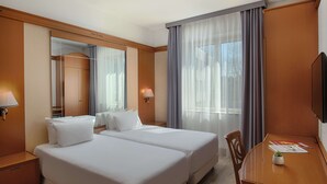A hotel room with two beds, a wooden desk, a TV, and a large window with curtains.