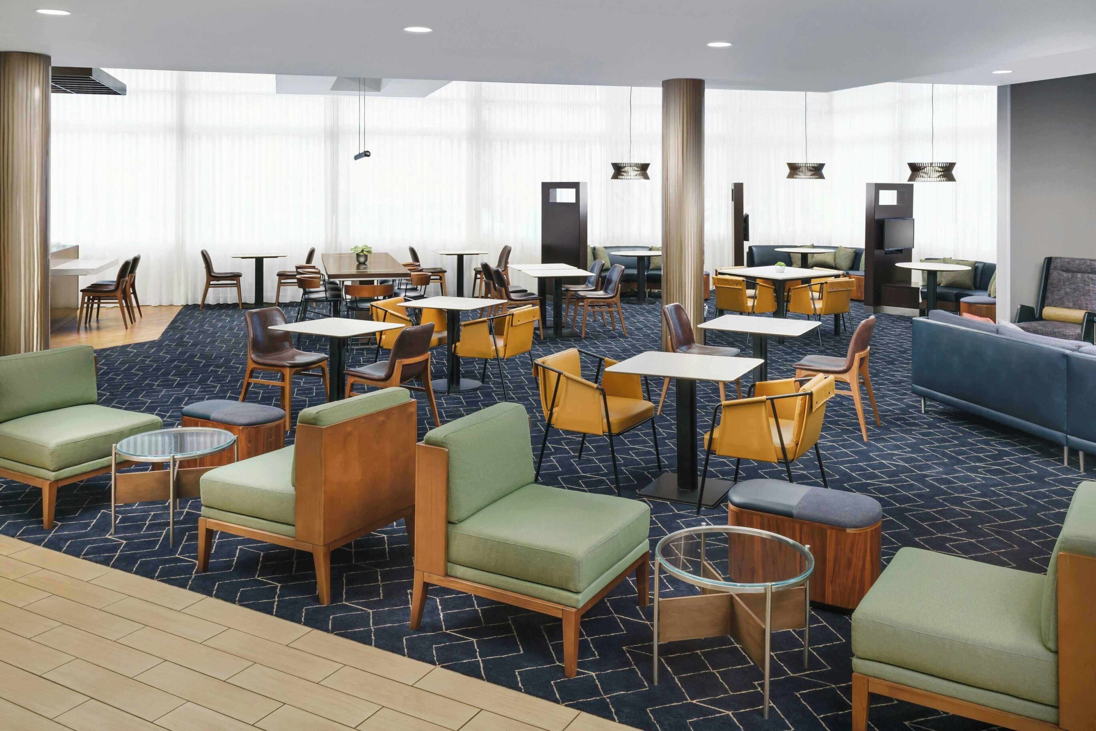 Un hall d’hôtel doté de plusieurs espaces de détente, comprenant des canapés, des fauteuils et des tables. De grandes fenêtres, ornées de rideaux légers, laissent entrer la lumière naturelle.
