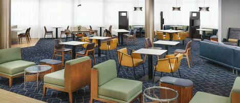 A hotel lobby with multiple seating areas, including sofas, chairs, and tables. There are large windows with sheer curtains, providing natural light.