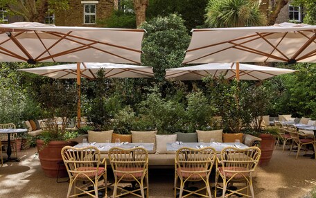 An outdoor seating area with wicker tables and chairs, surrounded by large umbrellas and potted plants.