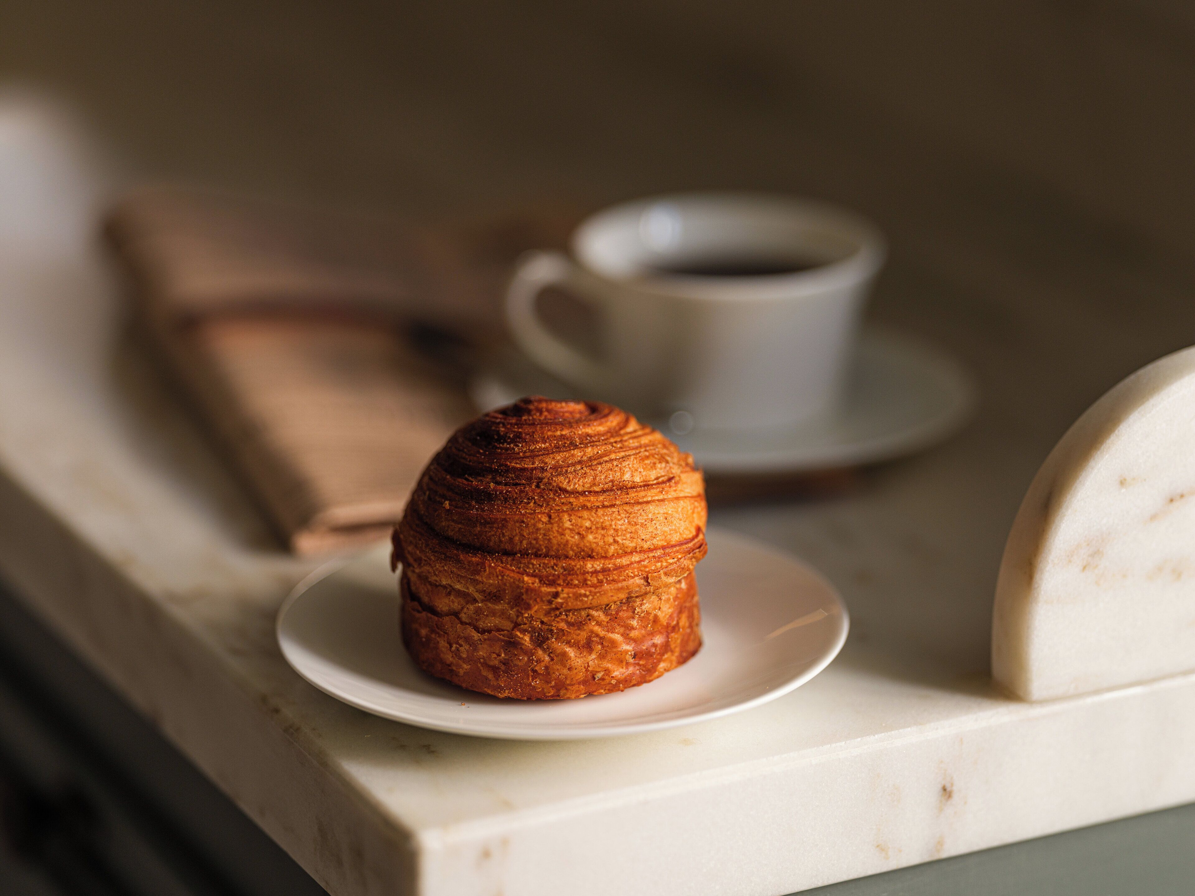 Ein Croissant auf einem weißen Teller, im Hintergrund eine Kaffeetasse.