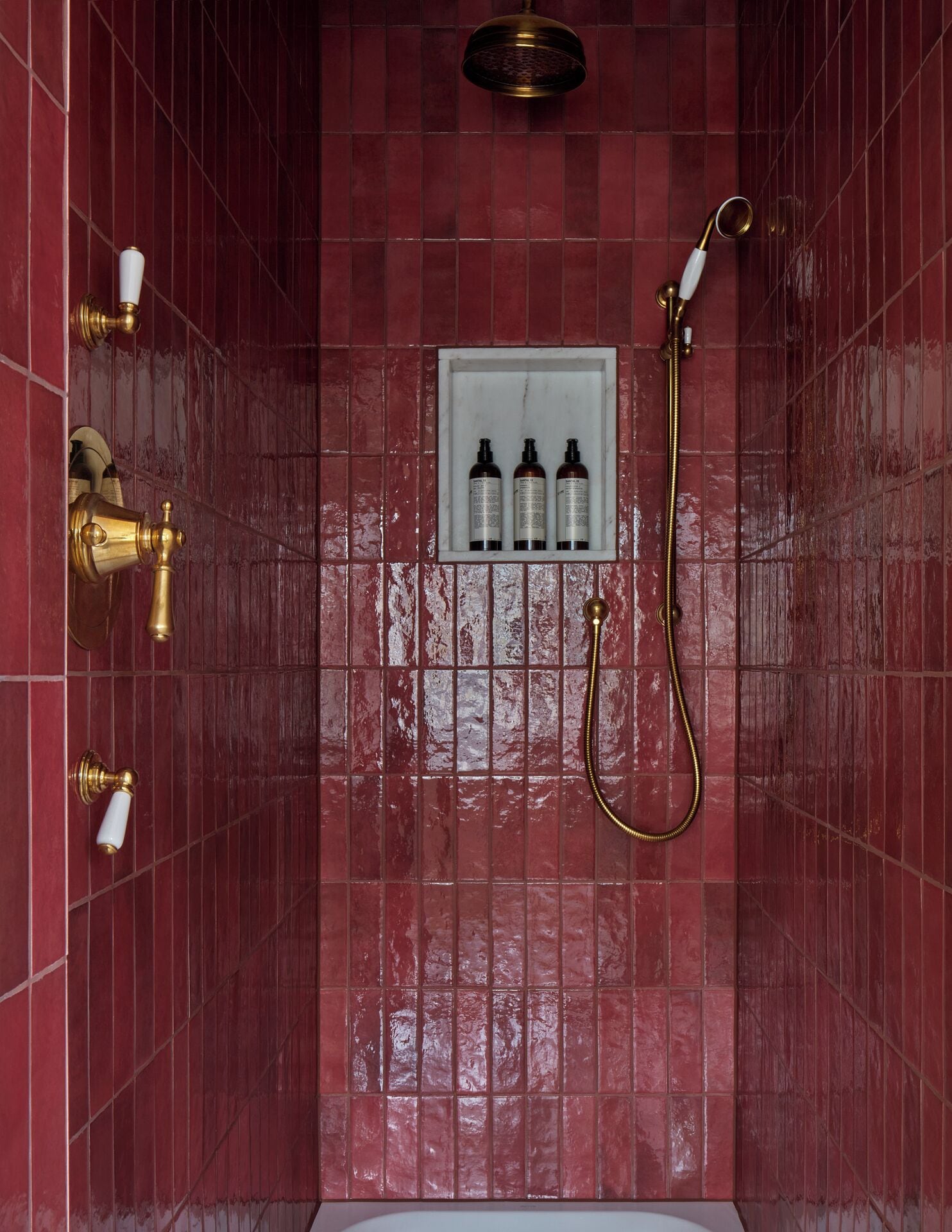 A bathroom with red tiled walls, a showerhead, and three bottles of shampoo in a white shelf.