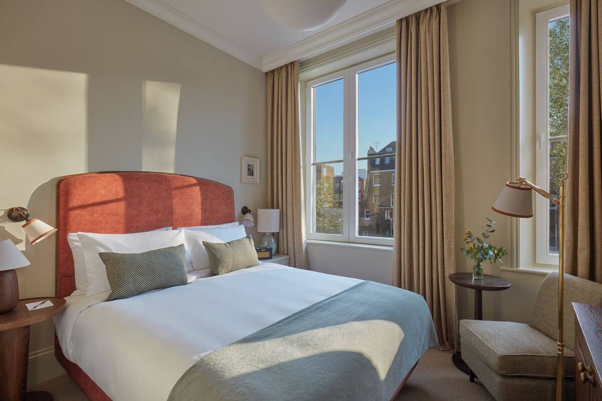 Ein Hotelzimmer mit einem großen Bett, einem Nachttisch mit Lampe, einem Sessel und einem Fenster mit Vorhängen.