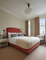 A hotel room with a large bed, a bedside table with a lamp, a chair, and a view of buildings outside the window.