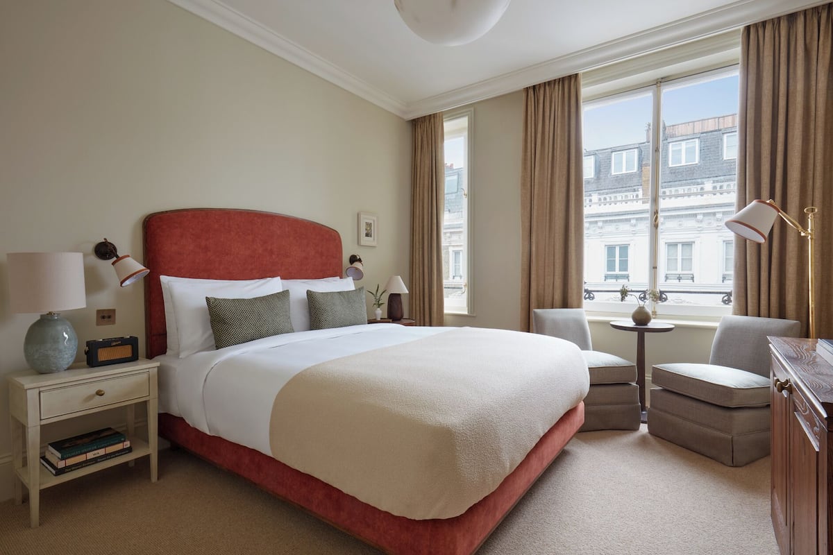 Ein Hotelzimmer mit einem großen Bett, einem Nachttisch mit Lampe, einem Sessel, einem kleinen Tisch und Blick auf die Stadt durch das Fenster.