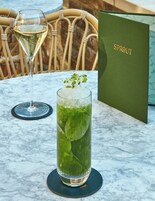 Ein hohes Glas mit einem grünen Getränk, Eiswürfeln und einem Spross als Garnitur auf einem Marmortisch. Außerdem sind ein Weinglas und eine Speisekarte zu sehen.