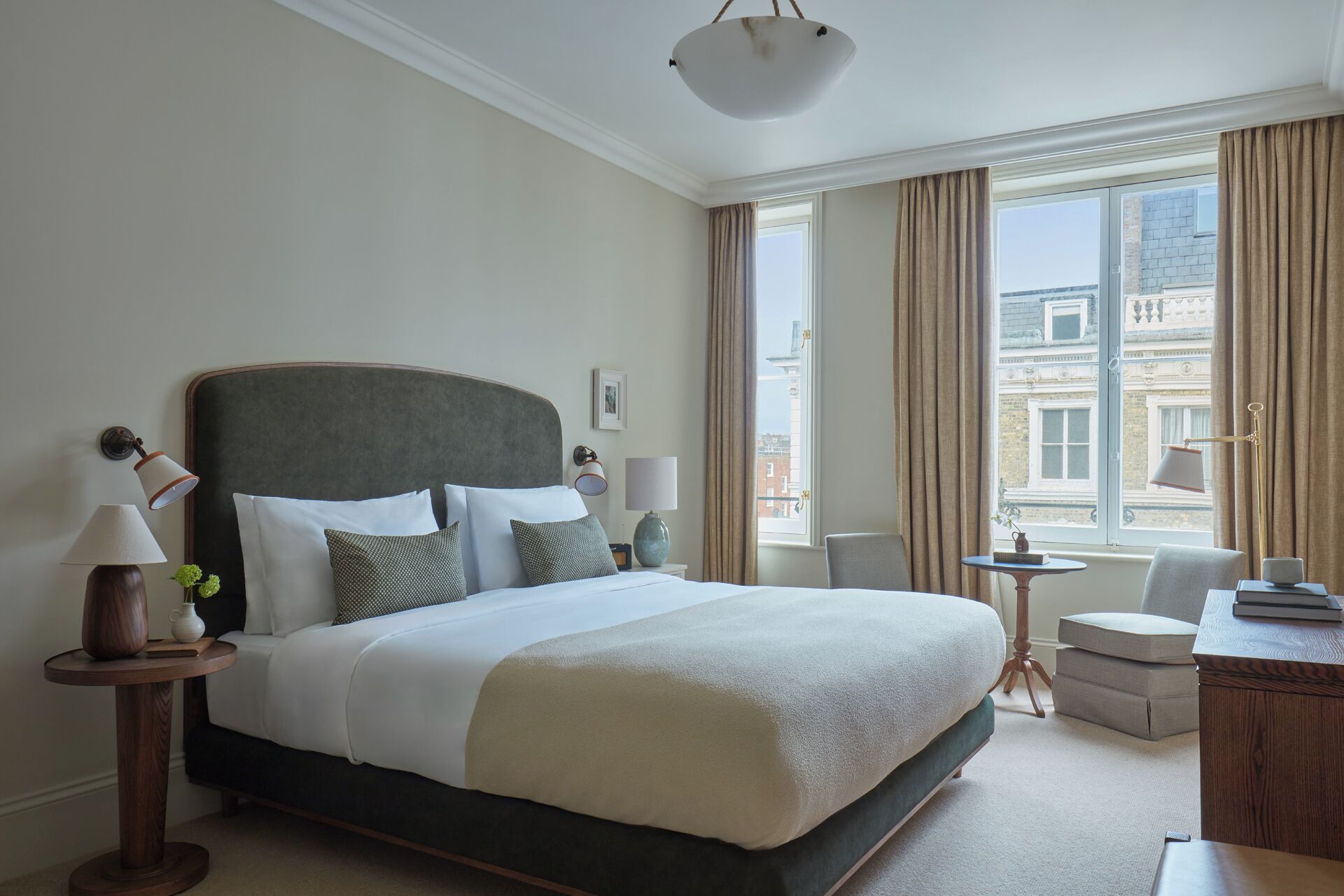 A hotel room with a large bed, bedside lamps, a small table, a chair, and a view of buildings through the window.