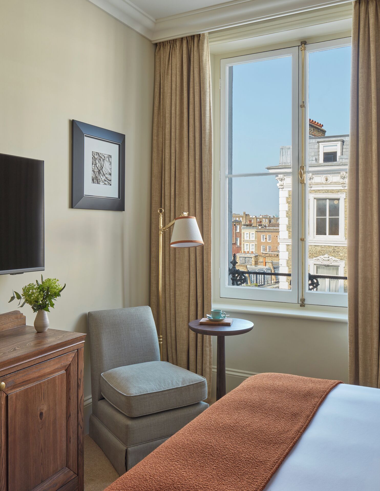 A hotel room with a bed, a chair, a small table with a cup and book, a lamp, a TV, and a window with a view of buildings.