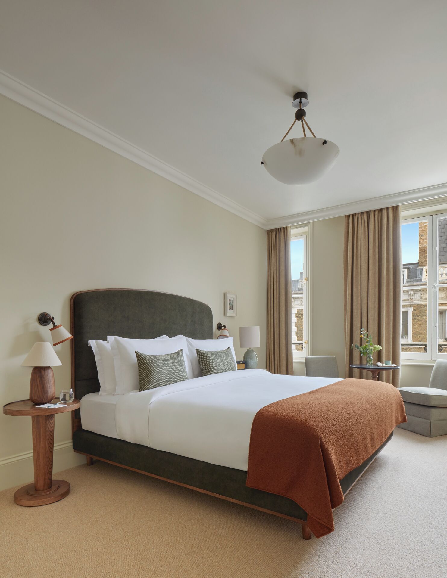 A hotel room with a large bed, a round wooden side table, a lamp, and a view of the city through the window.