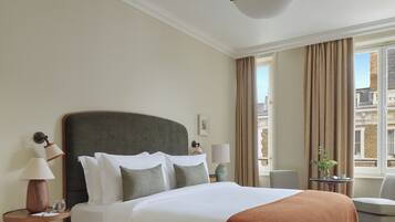 A hotel room with a large bed, a round wooden side table, a lamp, and a view of the city through the window.