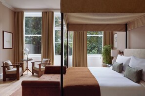 Una habitación de hotel con una cama grande, una zona de estar con una silla y una mesa, y ventanas amplias con cortinas.