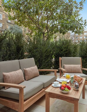 Un espacio al aire libre con sofás y sillones, una mesa con alimentos para el desayuno, y vista a un edificio en el fondo.