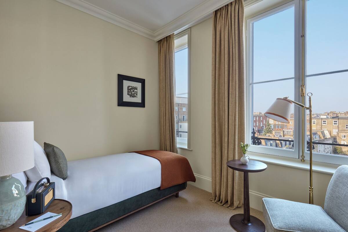 Ein Hotelzimmer mit einem Bett, einem Schreibtisch mit Lampe, einem Sessel, einem Fenster mit Vorhängen und Blick auf Gebäude.