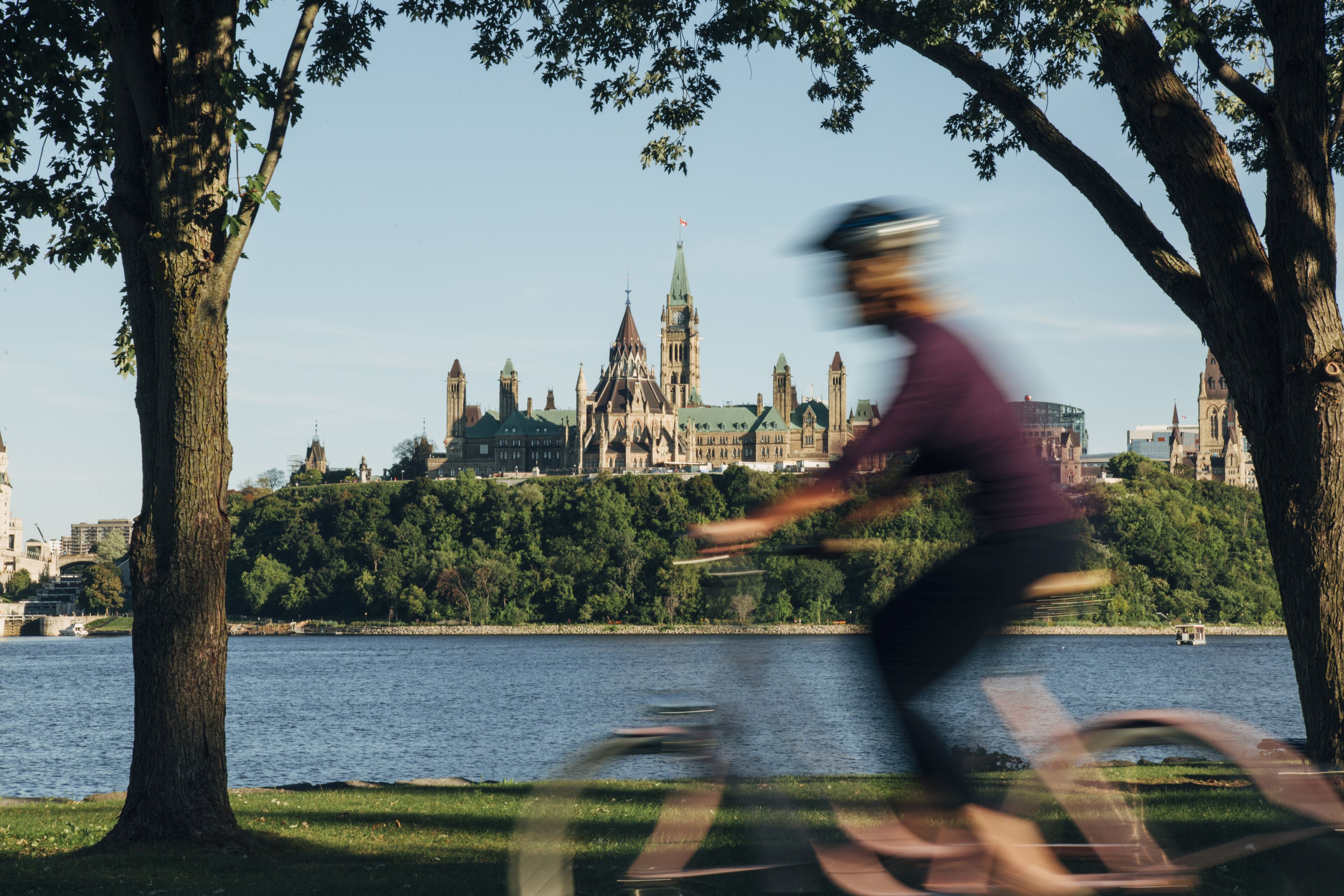 Un cycliste passe devant un bâtiment historique doté d’une grande tour horlogère et de flèches, entouré d’arbres et bordé par un plan d’eau.