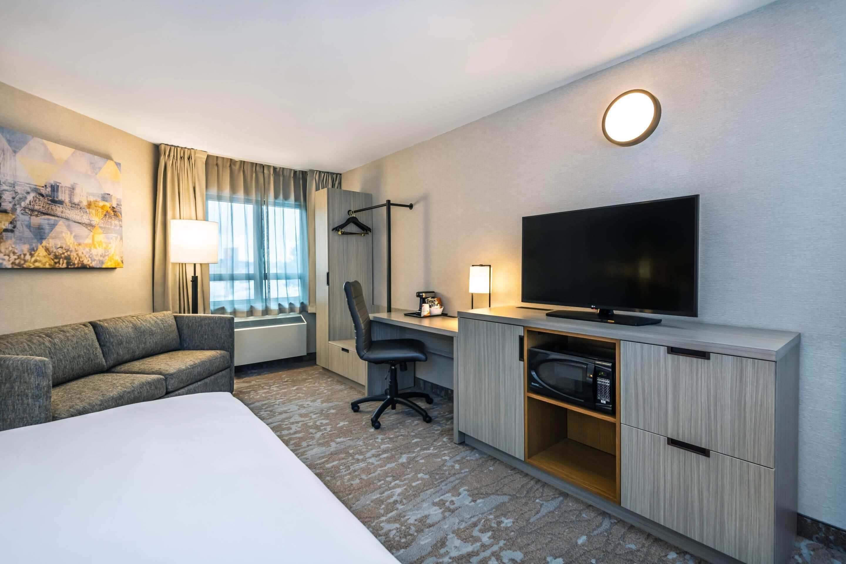 Ein Hotelzimmer mit Bett, Schreibtisch mit Computer, Fernseher, Sessel, Lampe und einem Bild an der Wand.