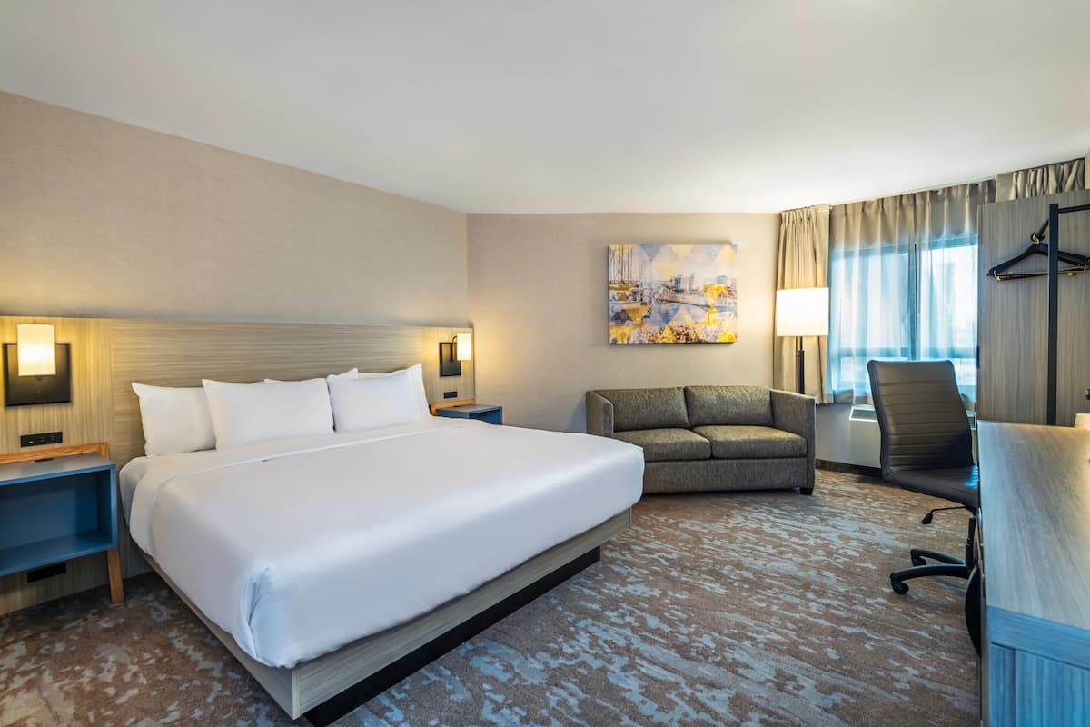 A hotel room with a large bed, a sofa, a desk, and a chair. There is a painting on the wall and a window with curtains.