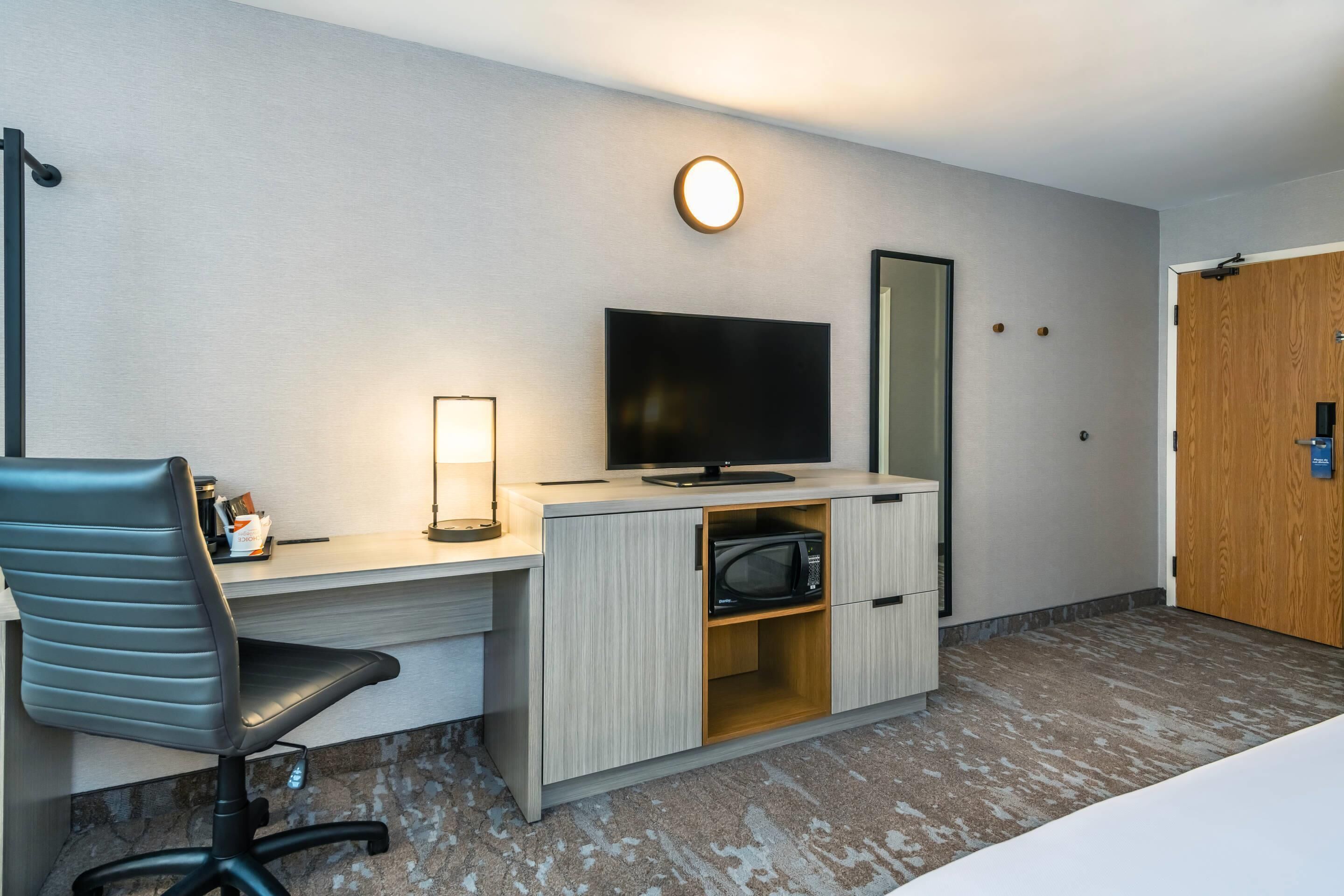 A hotel room with a desk, a chair, a flat-screen TV, a microwave, and a wooden door.