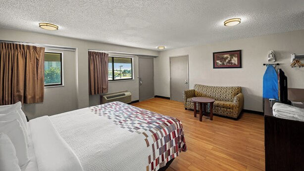 A hotel room with a bed, a sofa, a small table, a TV, and a window with curtains. - Red Roof Inn Dallas - DFW Airport North (Irving)