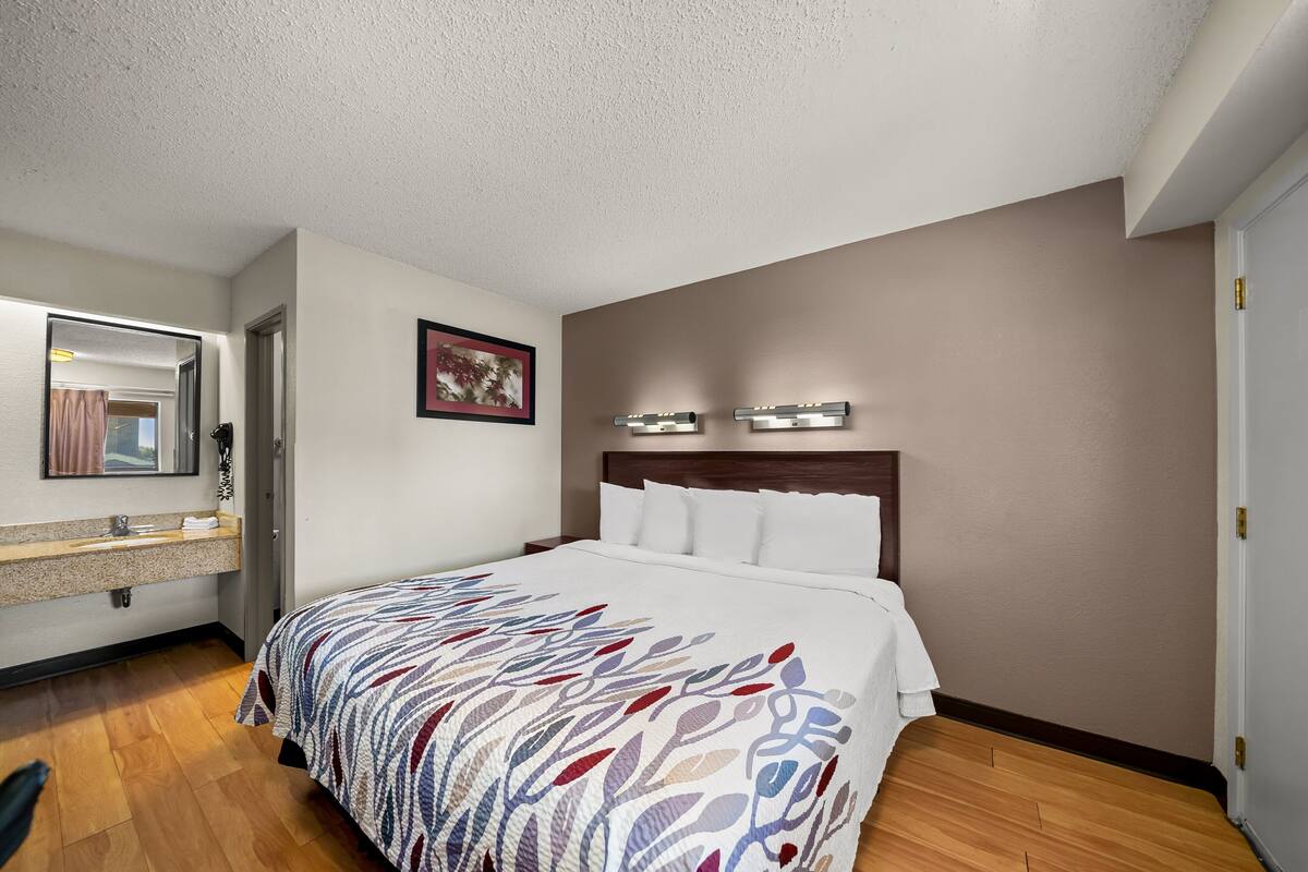 A hotel room with a bed, a vanity with a mirror, and a wall-mounted light fixture.