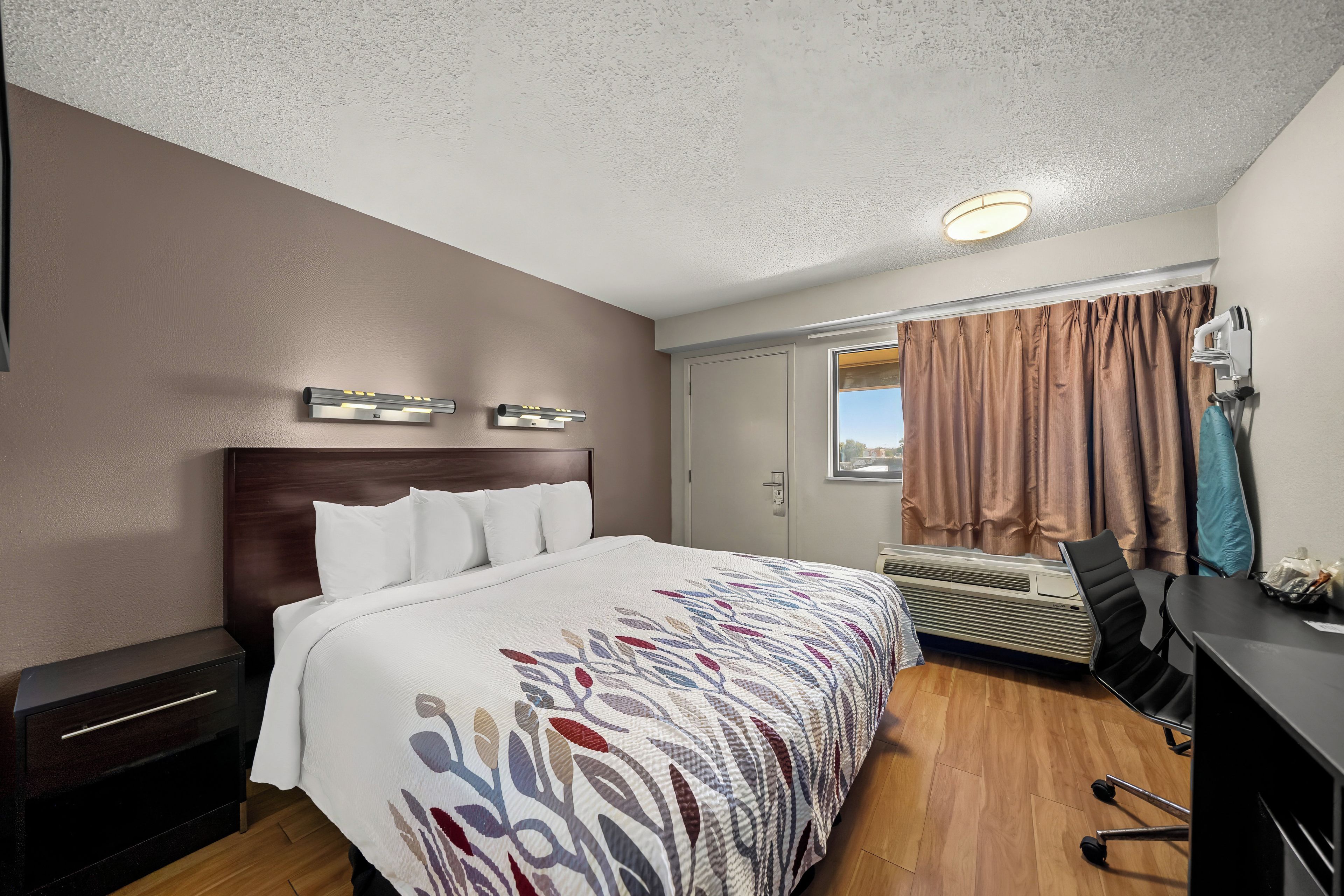 Ett hotellrum med en säng, ett skrivbord, en stol, en luftkonditioneringsanläggning och ett fönster med gardiner.