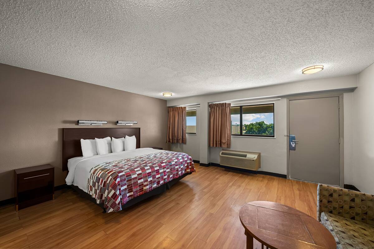 A hotel room with a large bed, a wooden floor, a small table, and a window with curtains.